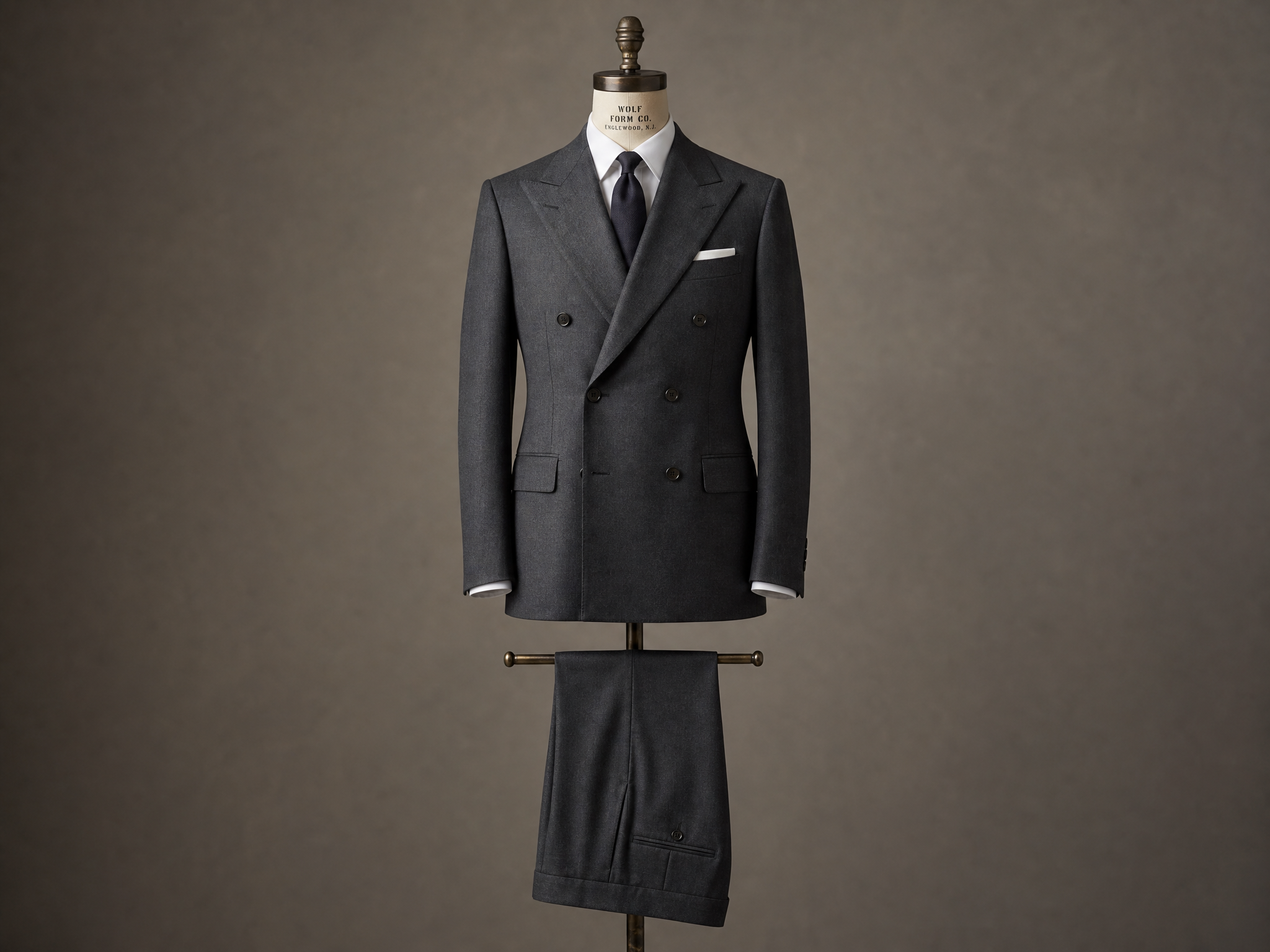 A finished charcoal double-breasted made-to-measure suit on a vintage Wolf Form Co. dressmaker's bust, against a warm taupe studio backdrop