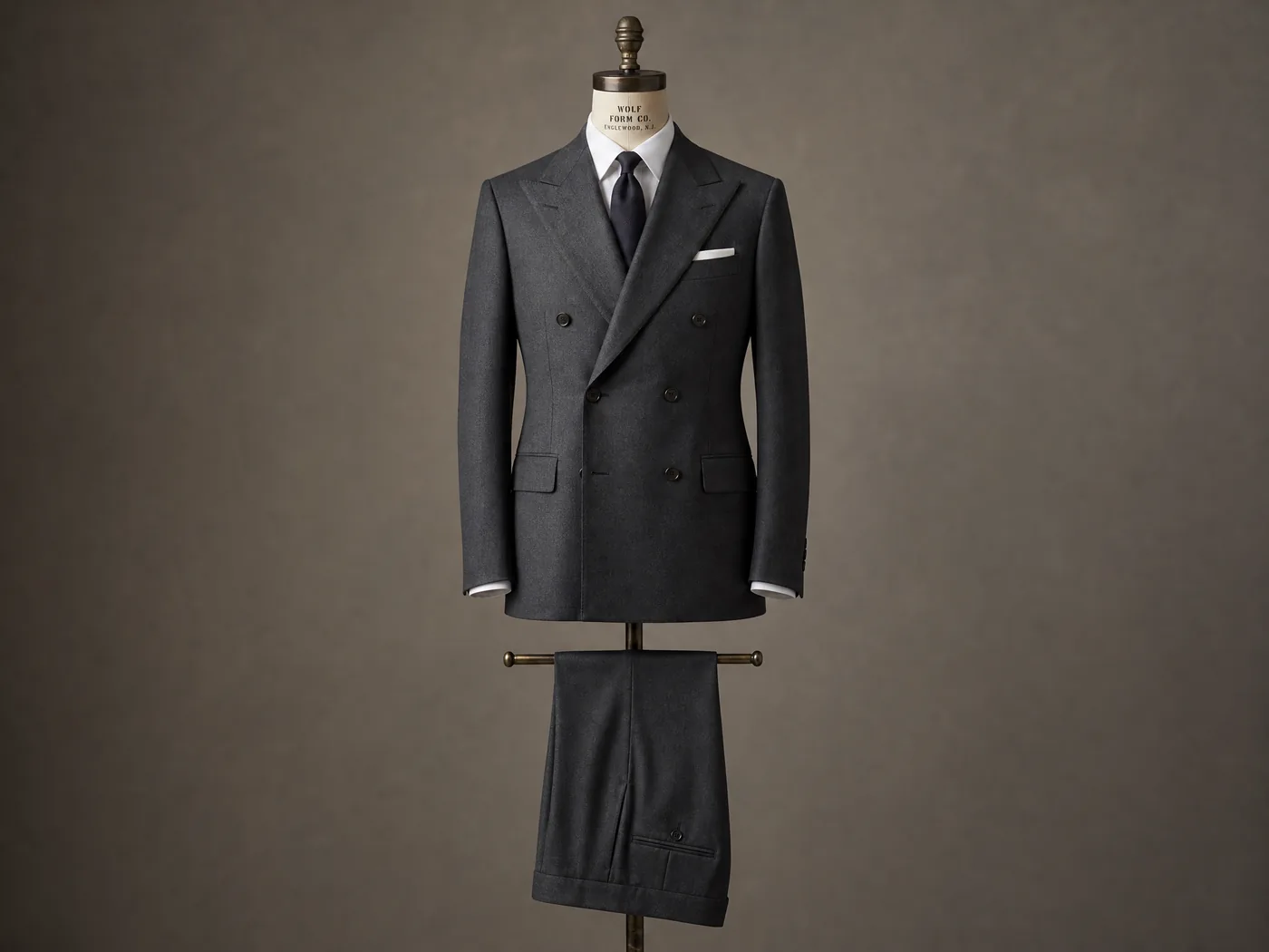 A finished charcoal double-breasted made-to-measure suit on a vintage Wolf Form Co. dressmaker's bust, against a warm taupe studio backdrop